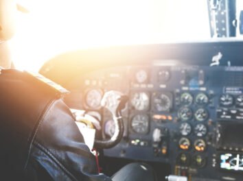 Caucasian aviator piloting airplane. Rear view of commercial pilot wearing headset and leather jacket sitting inside cockpit in front of dashboard with numerous buttons. Blurred shot, flare sun
